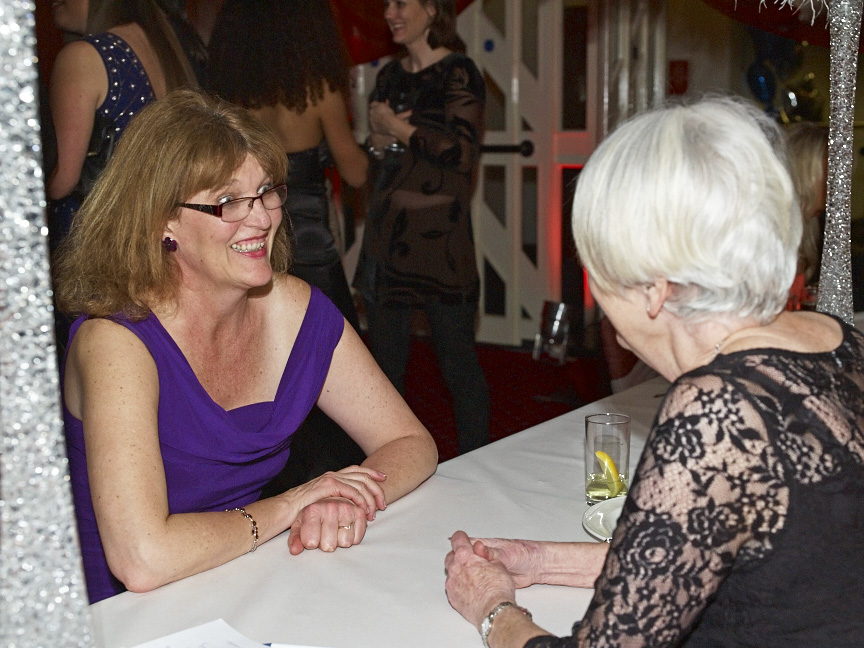 Two women in formal wear chatting at a white table with drinks and people in background at Healix Christmas event
