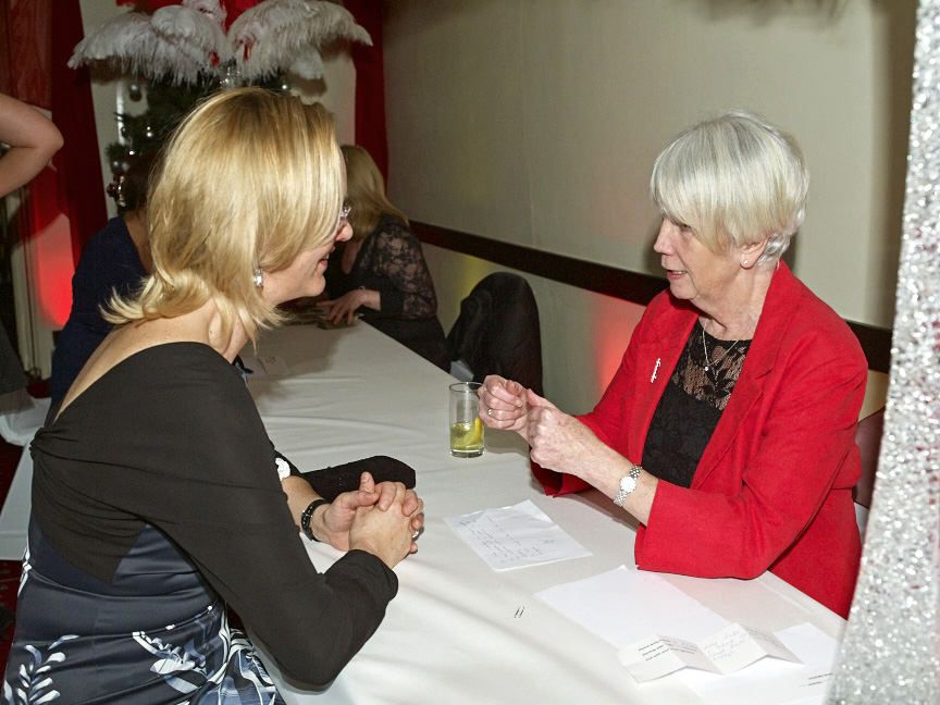 Two women in formal wear talking at a white table with papers and drinks