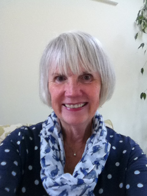 A smiling woman with short gray hair and bangs wearing a blue polka dot shirt and scarf indoors