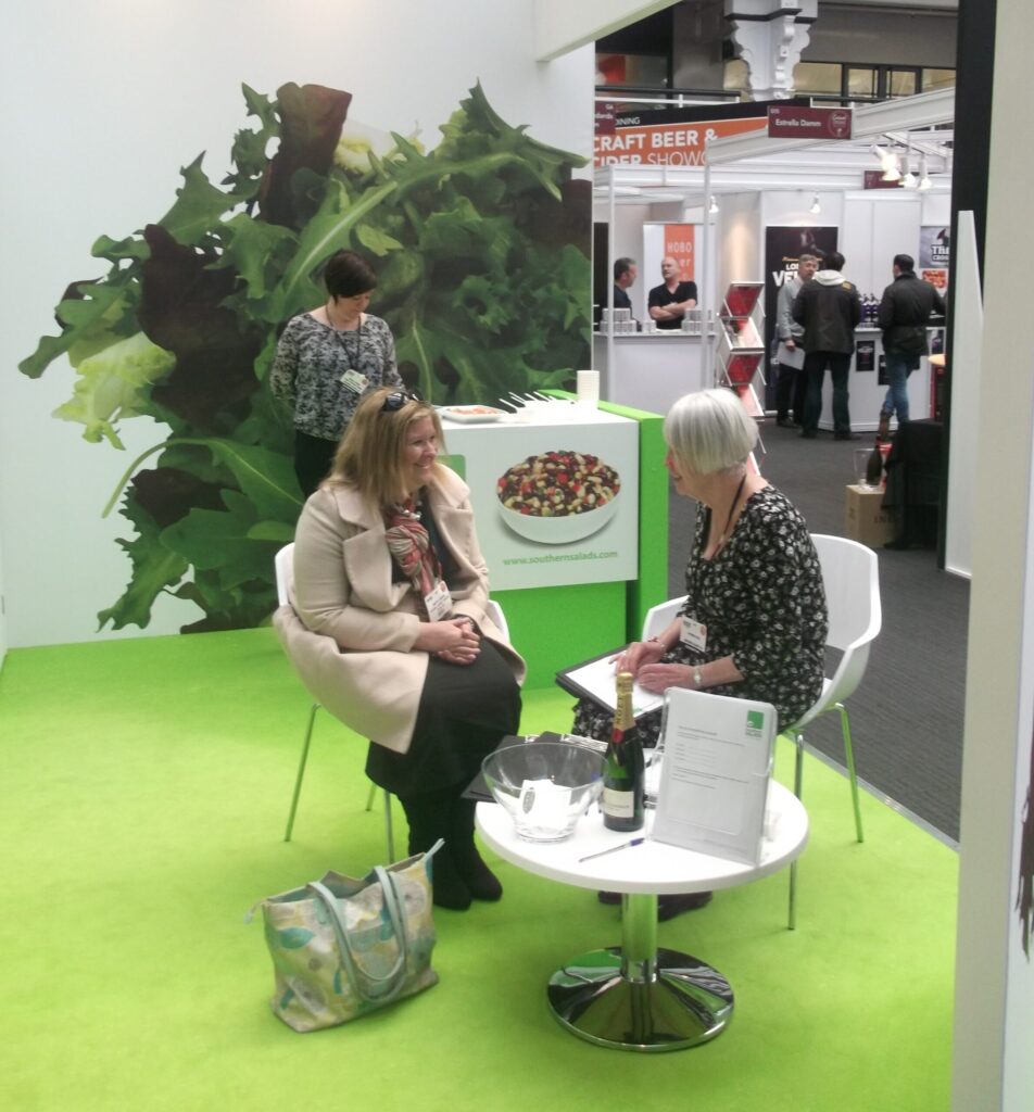Two women sit on chairs at a green-floored booth with a salad display, chatting and smiling, near a craft beer area.